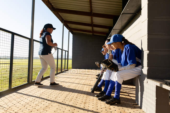 Grupo diverso de jogadoras de beisebol sentadas no banco, ouvindo treinadoras antes do jogo. time de beisebol feminino, treinamento esportivo e táticas de jogo. — Fotografia de Stock