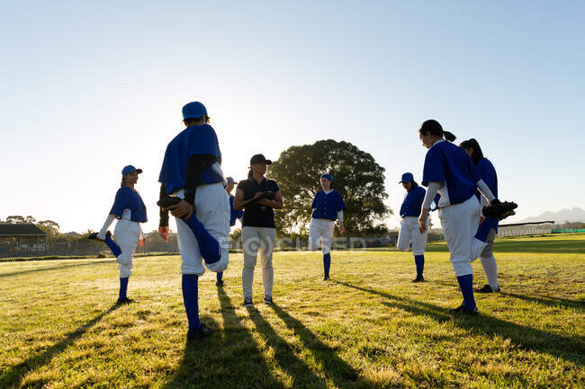 Groupe diversifié de joueuses de baseball avec entraîneur, se réchauffant sur le terrain, debout, jambes tendues. équipe féminine de baseball, entraînement sportif, convivialité et engagement. — Photo de stock