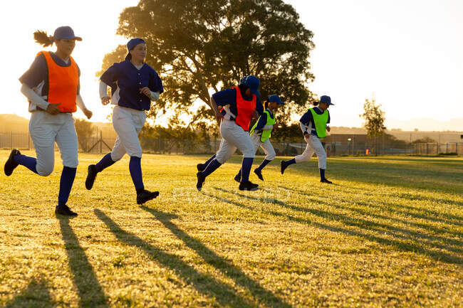 Diverso grupo de jugadoras de béisbol que se calientan en el campo al amanecer, corriendo. equipo femenino de béisbol, entrenamiento deportivo, unión y compromiso. - foto de stock