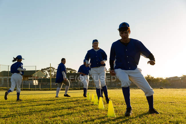 Diverse group of female baseball players warming up on field at sunrise, running slalom around cones. female baseball team, sports training and game tactics. — Stock Photo