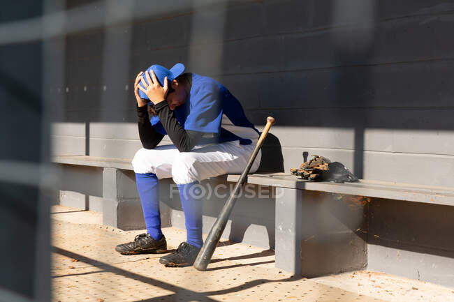 Mixed race female baseball player sitting on bench with head in hands during game. female baseball team, sports training and game tactics. — Stock Photo