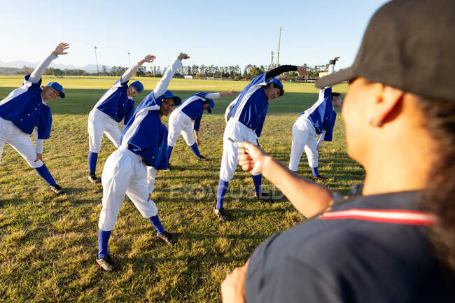 Vielfältige Gruppe von Baseballspielerinnen mit Trainer, die sich auf dem Feld aufwärmen und sich von der Hüfte strecken. Baseballmannschaft, Sporttraining, Zusammenhalt und Engagement. — Stockfoto