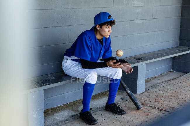 Mixed race female baseball player sitting on bench throwing ball, waiting to play during game. female baseball team, sports training and game tactics. — Stock Photo