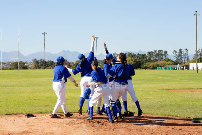 Разнообразная группа счастливых бейсболисток, празднующих на солнечном бейсбольном поле после игры. женская бейсбольная команда, спортивные тренировки, сплоченность и приверженность. — стоковое фото