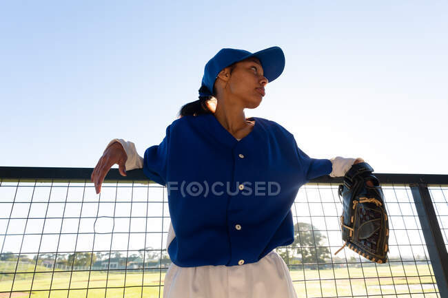 Giocatrice mista di baseball che indossa un guanto, appoggiata alla recinzione al sole prima della partita. squadra di baseball femminile, allenamento sportivo e tattica di gioco. — Foto stock