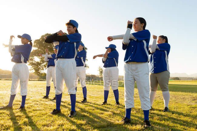Diverso gruppo di giocatrici di baseball che si scaldano sul campo soleggiato, allungando le braccia. squadra di baseball femminile, allenamento sportivo, solidarietà e impegno. — Foto stock