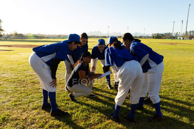 Un variegato gruppo di giocatrici di baseball che si stringono intorno all'allenatore accovacciato sul campo. squadra di baseball femminile, allenamento sportivo e tattica di gioco. — Foto stock