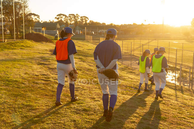 Groupe diversifié de joueuses de baseball marchant sur le terrain au lever du soleil pour s'entraîner. équipe féminine de baseball, entraînement sportif, convivialité et engagement. — Photo de stock