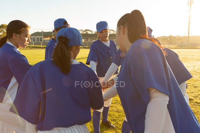 Diverse Baseballspielerinnen wärmen sich bei Sonnenaufgang auf dem Feld auf und stapeln Hände. Baseballmannschaft, Sporttraining, Zusammenhalt und Engagement. — Stockfoto
