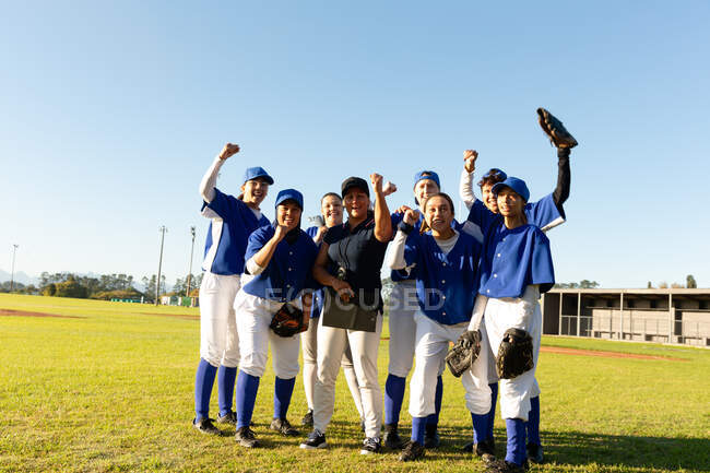 Портрет разнообразной группы женщин-бейсболистов и тренеров, стоящих и болеющих на солнечном поле. женская бейсбольная команда, спортивные тренировки, сплоченность и приверженность. — стоковое фото