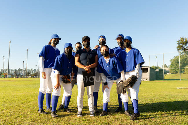 Porträt einer bunt gemischten Gruppe von Baseballspielerinnen und Trainerinnen mit Gesichtsmasken, die auf einem sonnigen Feld stehen. Baseballteam der Frauen, Sporttraining, Zusammenhalt und Engagement während der 19 Jahre währenden Pandemie. — Stockfoto