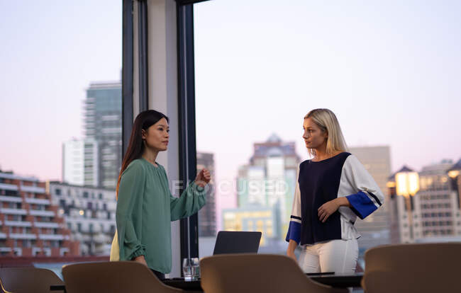 Zwei unterschiedliche Kolleginnen aus der Wirtschaft, die sich unterhalten und Laptop benutzen. Arbeit in einem modernen Büro. — Stockfoto