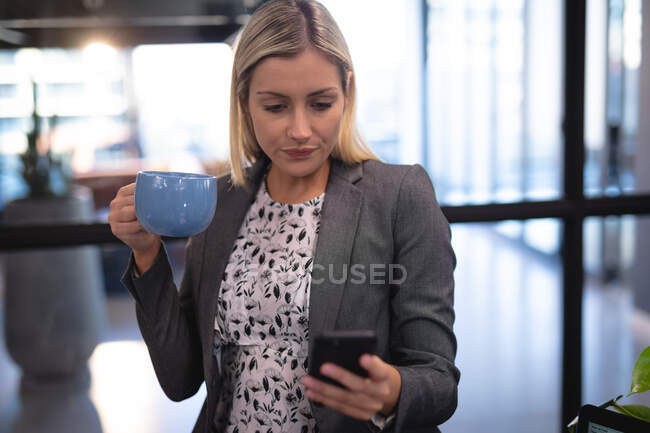 Caucasian businesswoman using smartphone and drinking mug of coffee. working in business at a modern office. — Stock Photo