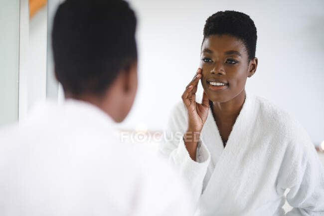 Mulher afro-americana sorridente na casa de banho a olhar para o espelho. estilo de vida doméstico, desfrutando de tempo de lazer em casa. — Fotografia de Stock