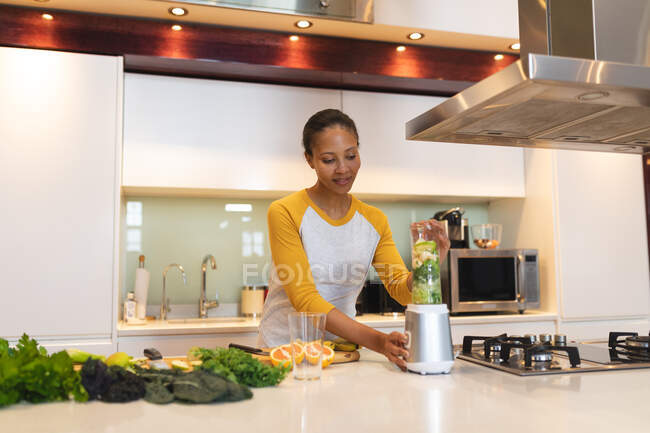 Sonriente mujer de raza mixta en la cocina preparando bebida saludable. estilo de vida doméstico, disfrutando del tiempo libre en casa. - foto de stock