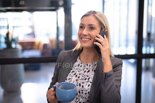 Кавказская деловая женщина, использующая смартфон и пьющая кружку кофе. работа в бизнесе в современном офисе. — стоковое фото