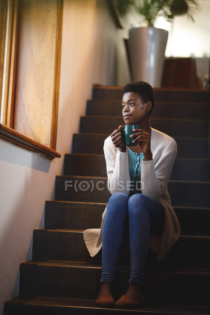 Femme afro-américaine souriante assise dans les escaliers, buvant du café. mode de vie domestique, profiter du temps libre à la maison. — Photo de stock