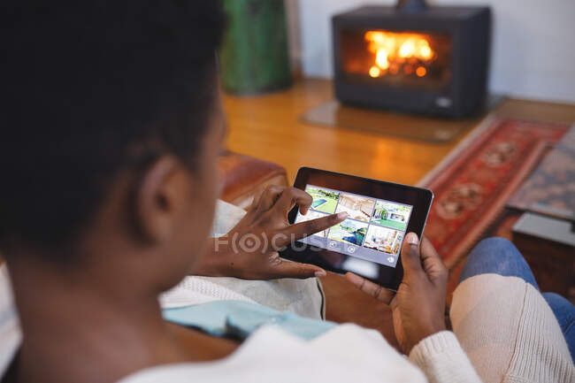Mujer afroamericana en la sala de estar sentada en un sofá y usando una tableta. estilo de vida doméstico, disfrutando del tiempo libre en casa. - foto de stock