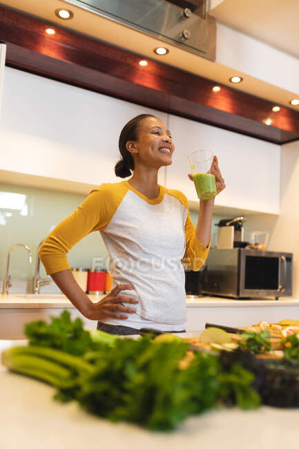 Mulher de raça mista sorridente na cozinha bebendo bebida saudável. estilo de vida doméstico, desfrutando de tempo de lazer em casa. — Fotografia de Stock