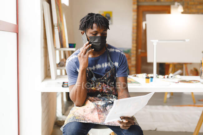 Afrikanisch-amerikanischer Maler mit Gesichtsmaske, der im Kunstatelier mit dem Smartphone spricht. Kreation und Inspiration in einem Malatelier während der Coronavirus-Pandemie 19. — Stockfoto