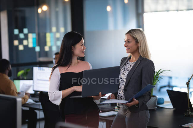 Dos compañeras de negocios diversas usando laptop y hablando. trabajar en un negocio en una oficina moderna. - foto de stock