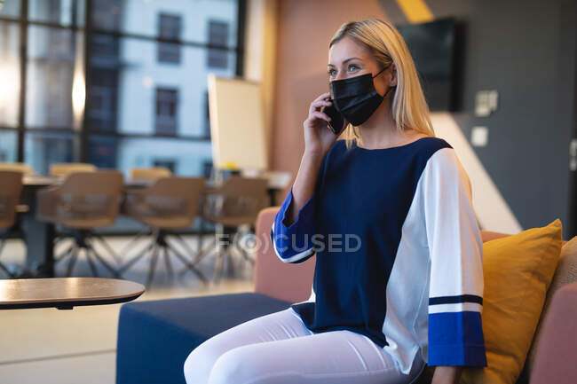Mulher de negócios caucasiana usando máscara facial e usando smartphone. trabalhando em negócios em um escritório moderno durante coronavírus covid 19 pandemia. — Fotografia de Stock
