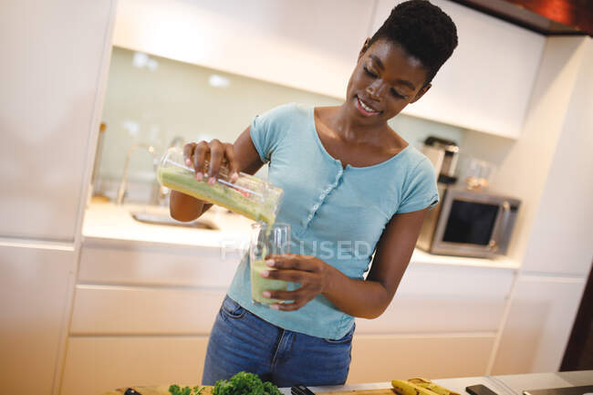 Femme afro-américaine souriante dans la cuisine préparant une boisson santé. mode de vie domestique, profiter du temps libre à la maison. — Photo de stock