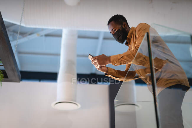 Empresário afro-americano usando máscara facial e usando smartphone. trabalhando em negócios em um escritório moderno durante coronavírus covid 19 pandemia. — Fotografia de Stock
