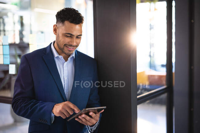 Hombre de negocios de raza mixta sonriente que usa tableta y chaqueta azul marino. trabajar en un negocio en una oficina moderna. - foto de stock