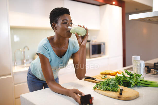 Mujer afroamericana en la cocina bebiendo bebidas saludables y usando un smartphone. estilo de vida doméstico, disfrutando del tiempo libre en casa. - foto de stock