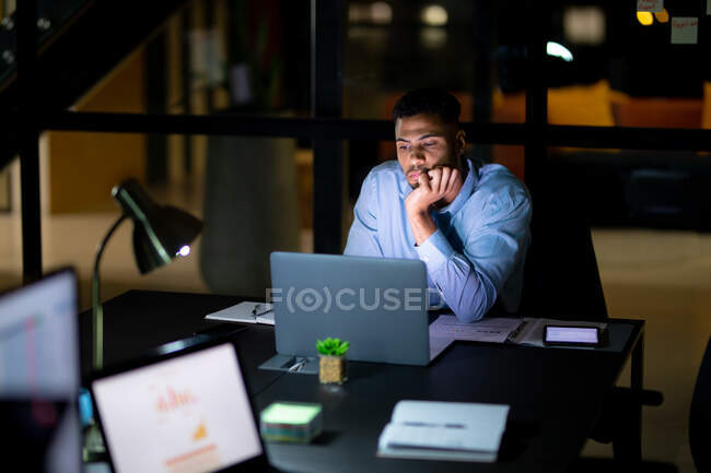 Бизнесмен смешанной расы работает по ночам, сидит за столом и пользуется ноутбуком. работать допоздна в современном офисе. — стоковое фото