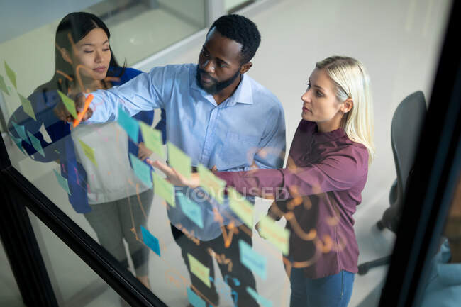 Diversos colegas de negocios masculinos y femeninos leyendo notas coloridas en la pared de vidrio. trabajar en un negocio en una oficina moderna. - foto de stock