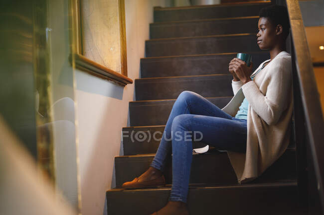 Femme afro-américaine souriante assise dans les escaliers, buvant du café. mode de vie domestique, profiter du temps libre à la maison. — Photo de stock