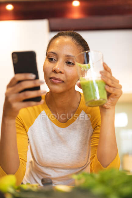 Усмішка змішаної раси жінка на кухні п'є алкогольний напій і користується смартфоном. Домашнє життя, дозвілля вдома. — стокове фото