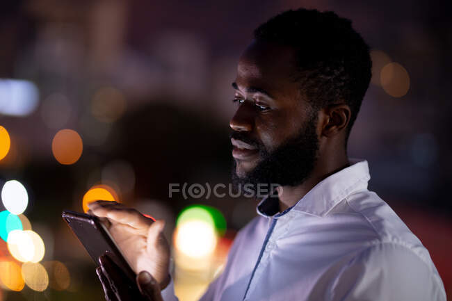 Hombre de negocios afroamericano que trabaja de noche con un smartphone. trabajar hasta tarde en los negocios en una oficina moderna. - foto de stock