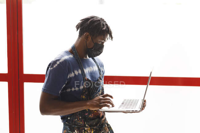 African american male painter wearing face mask using laptop in art studio. creation and inspiration at an artists painting studio during coronavirus covid 19 pandemic. — Stock Photo