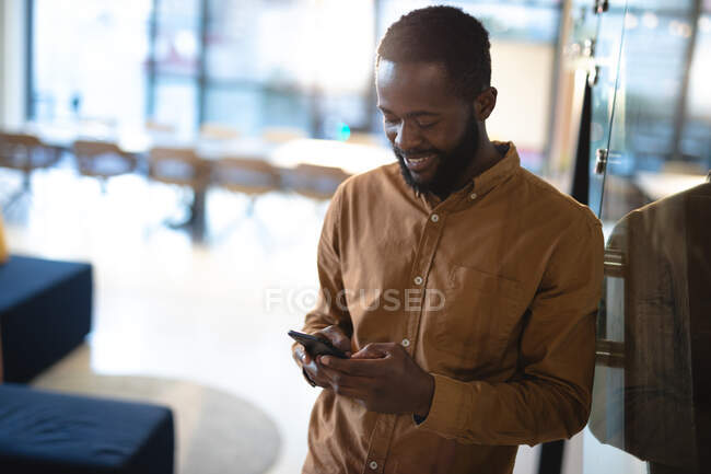 Un uomo d'affari afroamericano sorridente che usa lo smartphone e indossa abiti formali. lavorare in azienda in un ufficio moderno. — Foto stock