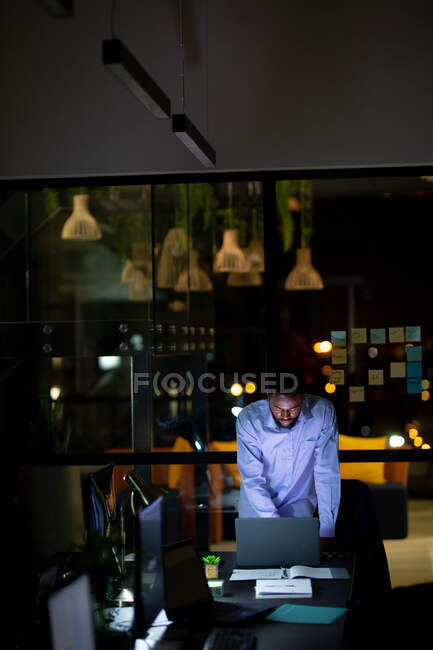 Африканский американский бизнесмен работает по ночам, стоит за столом и пользуется ноутбуком. работать допоздна в современном офисе. — стоковое фото