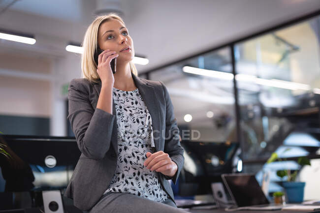 Donna d'affari caucasica seduta alla scrivania e che parla su smartphone. lavorare in azienda in un ufficio moderno. — Foto stock