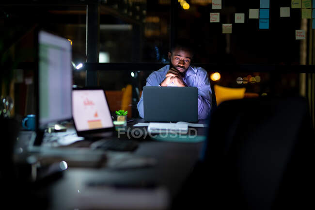 Hombre de negocios afroamericano trabajando de noche, sentado en el escritorio y usando un portátil. trabajar hasta tarde en los negocios en una oficina moderna. - foto de stock