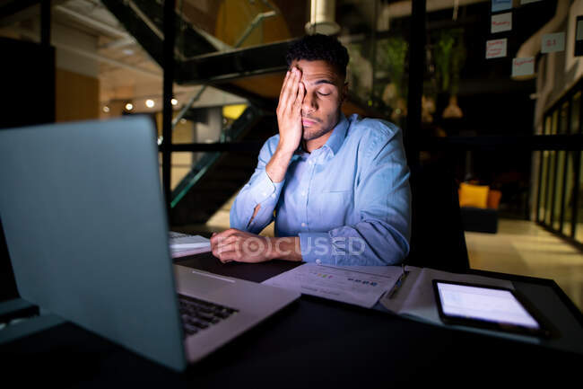 Empresario de carrera mixta trabajando de noche, sentado en el escritorio y usando un portátil. trabajar hasta tarde en los negocios en una oficina moderna. - foto de stock