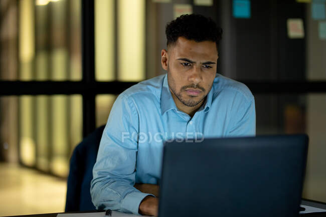 Empresario de carrera mixta que trabaja de noche usando laptop. trabajar hasta tarde en los negocios en una oficina moderna. - foto de stock