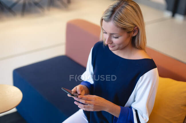 Kaukasische Geschäftsfrau, die auf der Couch sitzt und ihr Smartphone benutzt. Arbeit in einem modernen Büro. — Stockfoto