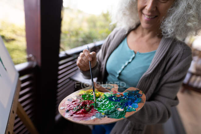 Happy senior caucasian woman standing on sunny balcony and painting. retirement lifestyle, spending time alone at home. — Stock Photo