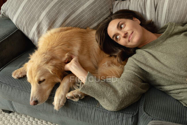 Femme caucasienne souriante dans le salon, allongée sur le canapé avec son chien de compagnie. mode de vie domestique, profiter du temps libre à la maison. — Photo de stock