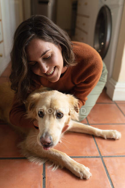 Lächelnde Kaukasierin in der Küche, die auf dem Boden sitzt und ihren Hund umarmt. häuslicher Lebensstil, Freizeit zu Hause genießen. — Stockfoto