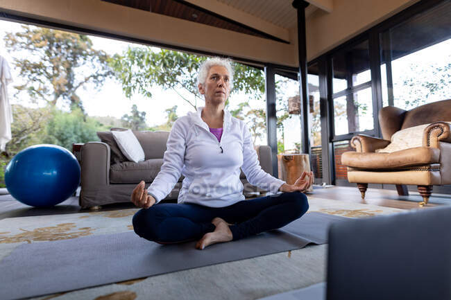 Senior caucasian woman in living room exercising and meditating, sitting on the floor. retirement lifestyle, spending time alone at home. — Stock Photo