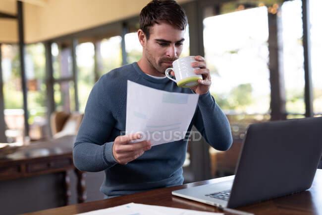 Homem caucasiano sentado à mesa e trabalhando remotamente usando laptop. trabalhando em casa no apartamento moderno. — Fotografia de Stock