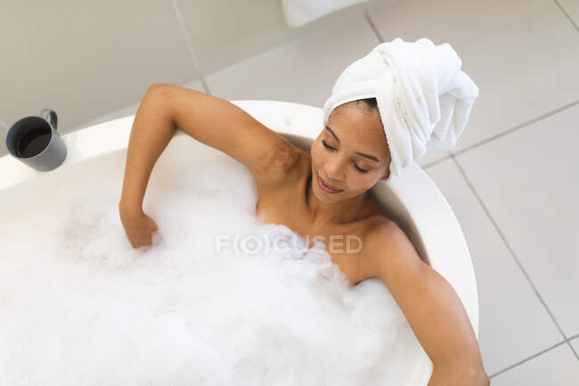 Mixed race woman in bathroom having a bath, relaxing with eyes closed. domestic lifestyle, enjoying self care leisure time at home. — Stock Photo