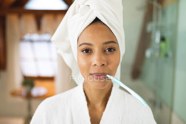 Retrato de mujer de raza mixta en el baño sosteniendo cepillo de dientes en la boca mirando a la cámara. - foto de stock